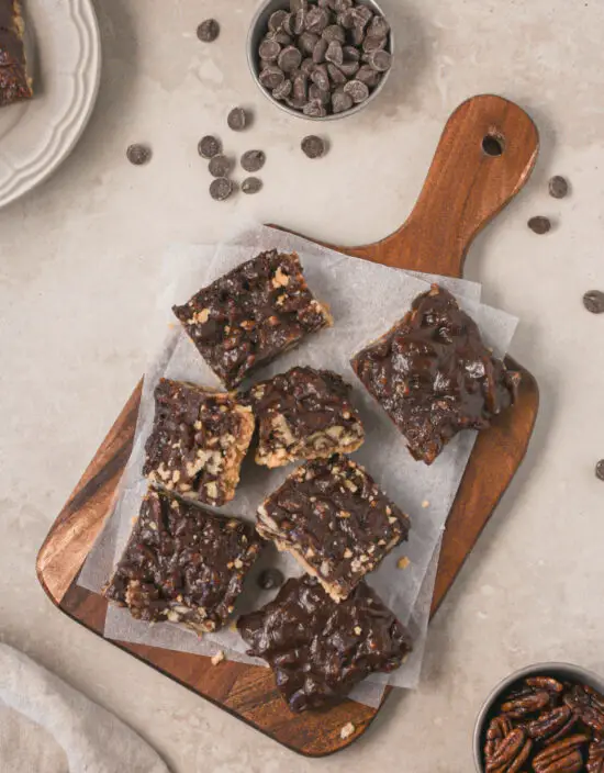 Wooden serving board piled with chocolate pecan bars on top with chocolate chips scattered around. 