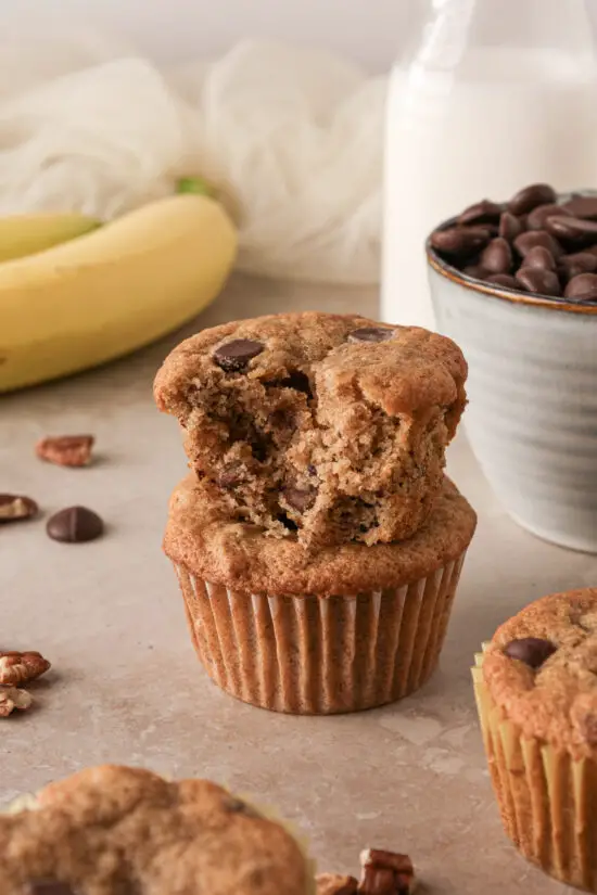 Two Banana Chocolate Chip Muffins stacked on top of each other with a banana and chocolate chips scattered around. 