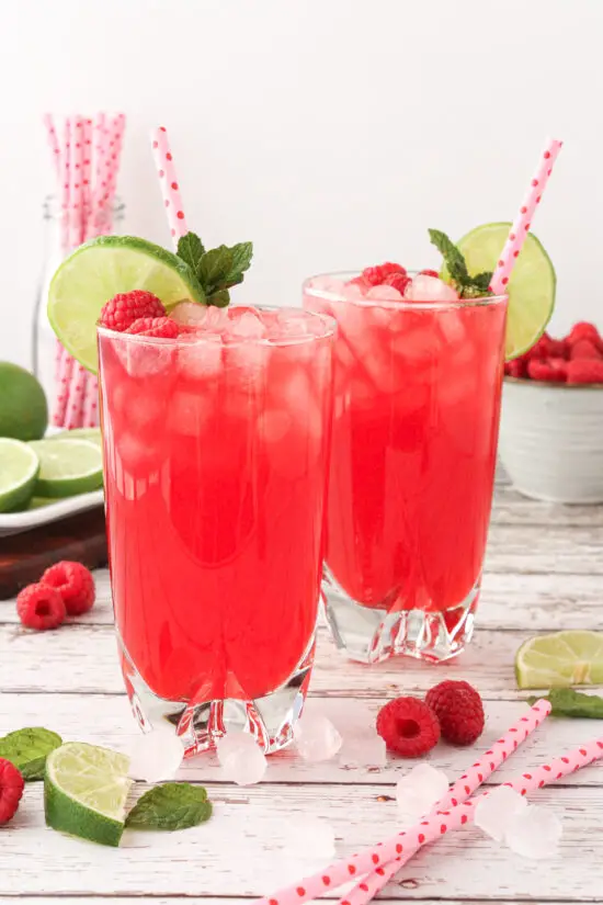 Two glasses filled with Raspberry Limeade, lime wedges, ice and straws in them. 