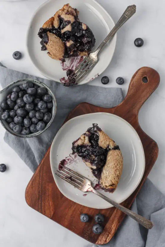 Two plates with slices of Blueberry Cobbler with forks and small bowl of fresh blueberries. 