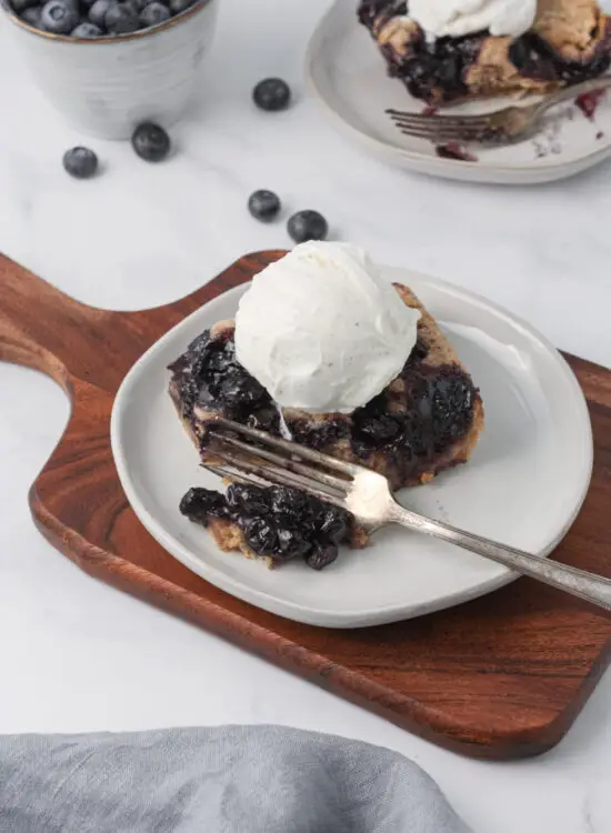 Slice of Blueberry Cobbler with scoop of vanilla ice cream on top on a plate with fork.