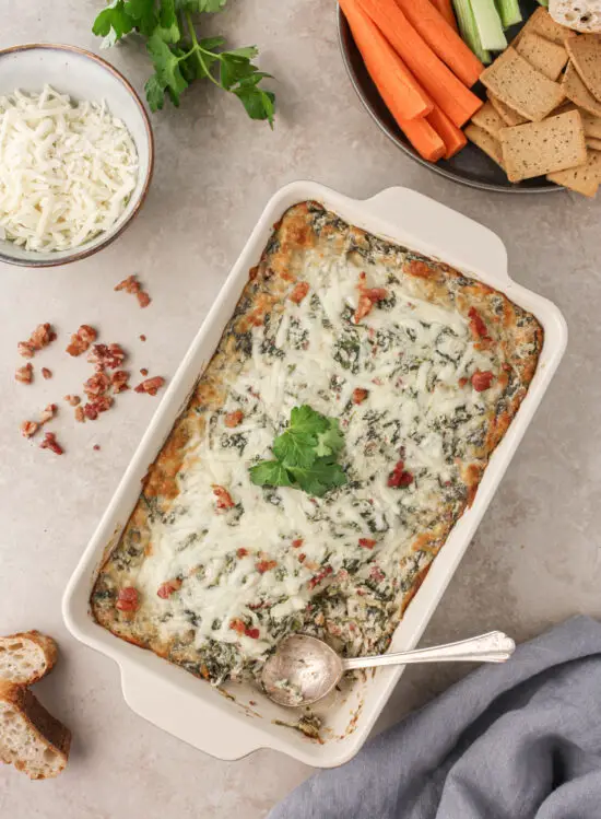 Baking pan filled with gooey Cheesy Bacon Spinach Dip and plate filled with fresh veggies and crackers for dipping.