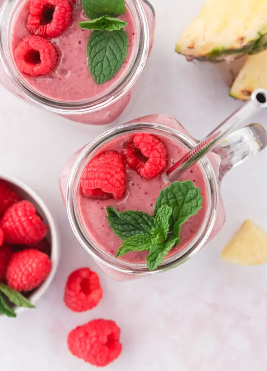 Two glass mugs filled with Pineapple Raspberry Protein Smoothie with fresh raspberries and mint on top.