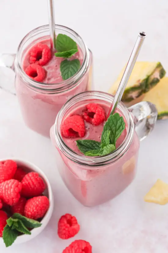 Two glass mugs filled with Pineapple Raspberry Protein Smoothie with straws, fresh mint, and raspberries on top.