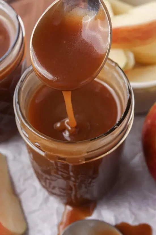 Spoon dipped in jar of Brown Sugar Caramel Sauce and sliced apples nearby. 